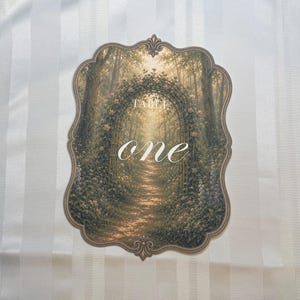 May include: A scalloped-edge table number card featuring a printed illustration of a sunlit forest path framed by a floral archway. The text "TABLE" appears in small capital letters above the large, white, cursive script word "one".