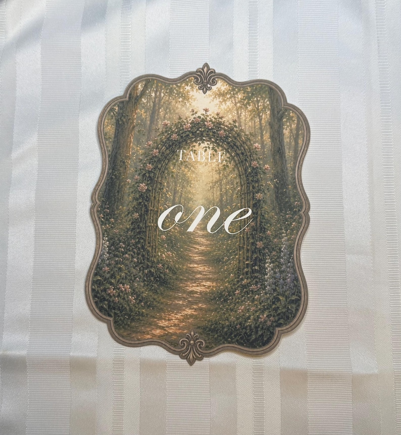 May include: A scalloped-edge table number card featuring a printed illustration of a sunlit forest path framed by a floral archway. The text "TABLE" appears in small capital letters above the large, white, cursive script word "one".