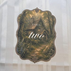 May include: A scalloped-edge table sign featuring a printed illustration of a stone cottage nestled in a lush, green forest setting. The text "TABLE" appears in a simple font above the large, white, cursive script word "two" in the center.