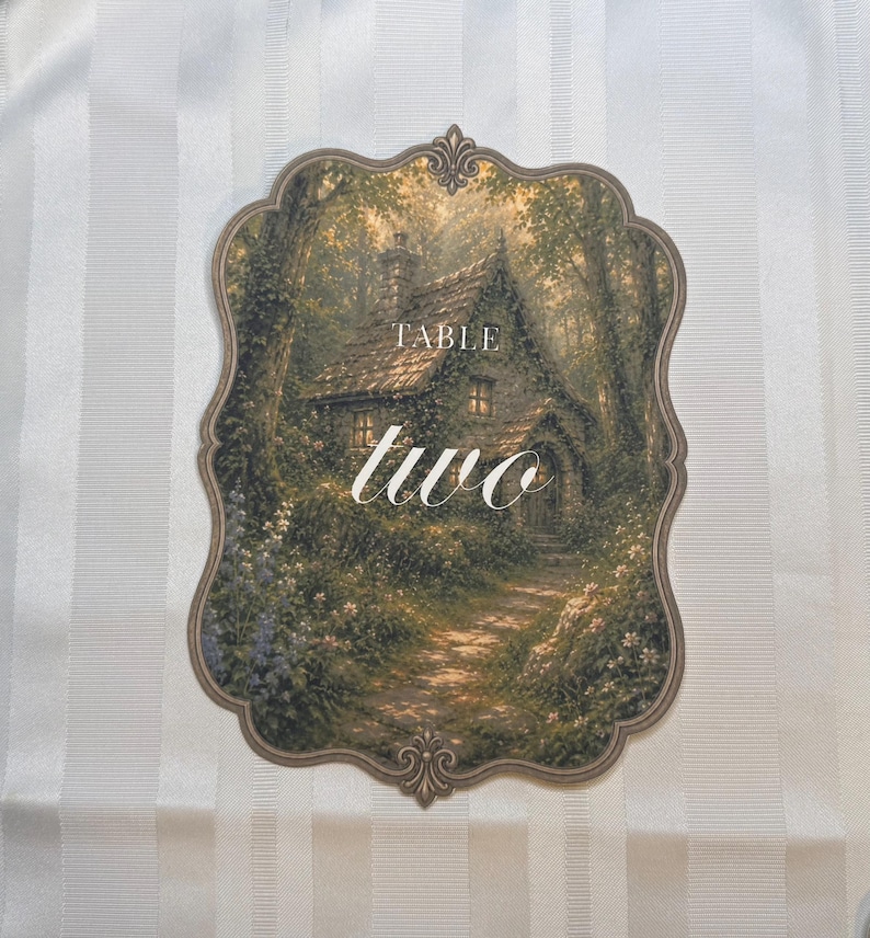 May include: A scalloped-edge table sign featuring a printed illustration of a stone cottage nestled in a lush, green forest setting. The text "TABLE" appears in a simple font above the large, white, cursive script word "two" in the center.