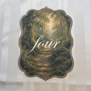 May include: A scalloped-edge table sign featuring a printed forest path scene with green trees and sunlight. The text "TABLE" appears in a small serif font above the large, white, cursive script word "four" centered on the card.
