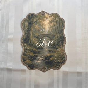 May include: A scalloped-edge table sign featuring a printed landscape illustration of a stone bridge over a flowing stream in a forest. The text "TABLE" appears in a simple font above the large, white, cursive script word "six" centered.