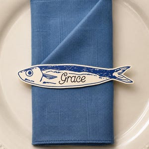 Op de afbeelding: Een blauw servet ligt op een wit bord, versierd met een visvormige naamkaart met de naam "Grace". Het visontwerp is blauw en wit, wat een nautische touch aan de tafelsetting geeft. Het bord heeft een parelrand.