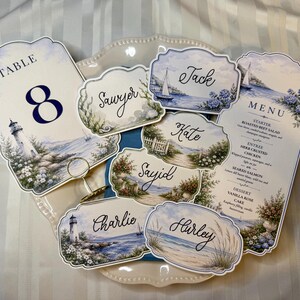 May include: A collection of wedding stationery featuring a coastal theme with lighthouse and ocean illustrations. The set includes a table number 8 card, a menu card, and several place cards with names like Jack, Sawyer, Kate, Sayid, Charlie, and Hurley.