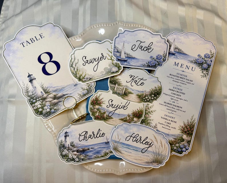May include: A collection of wedding stationery featuring a coastal theme with lighthouse and ocean illustrations. The set includes a table number 8 card, a menu card, and several place cards with names like Jack, Sawyer, Kate, Sayid, Charlie, and Hurley.