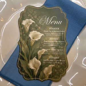 May include: A scalloped-edge menu card featuring a dark green background with white calla lily illustrations. The text reads Menu, Starter Fresh Fig & Burrata Salad, Entree Herb Grilled Filet Mignon or Lemon Thyme Chicken, and Dessert Vanilla Bean Creme Brulee.