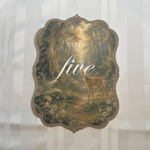 May include: A decorative table number card with a scalloped edge, featuring a printed forest scene with a small deer standing on a path. The text "TABLE five" is printed in white in the center of the card, which is set against a white striped background.
