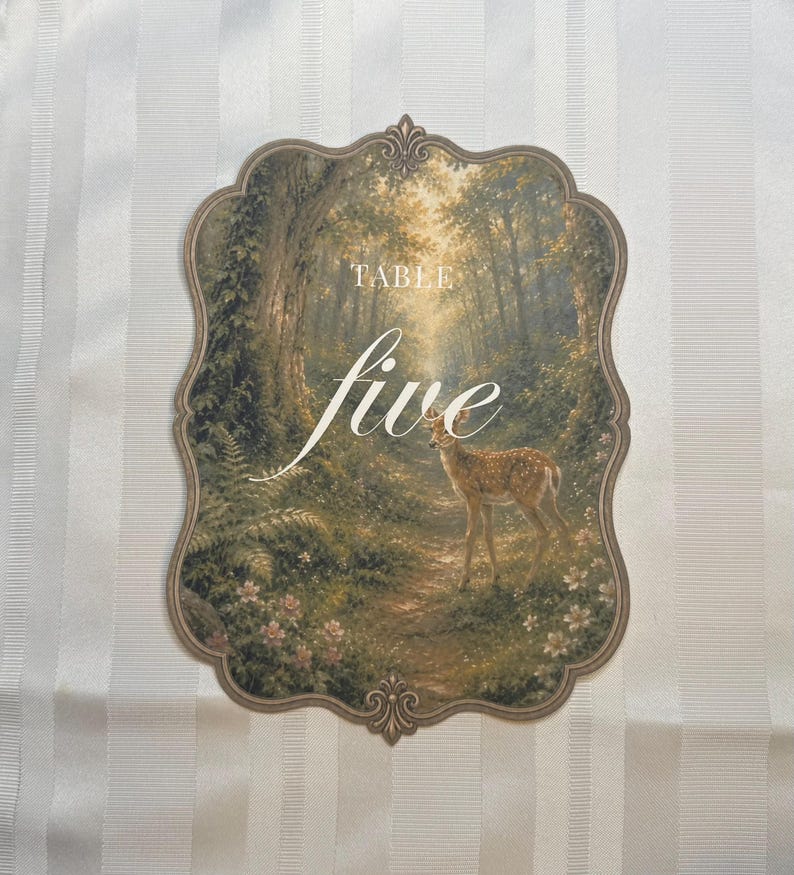 May include: A decorative table number card with a scalloped edge, featuring a printed forest scene with a small deer standing on a path. The text "TABLE five" is printed in white in the center of the card, which is set against a white striped background.