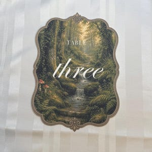 May include: A scalloped-edge table number card featuring a forest landscape with a stream, mossy rocks, and mushrooms. The text "TABLE" appears in a serif font above the word "three" written in a white cursive script centered on the card.