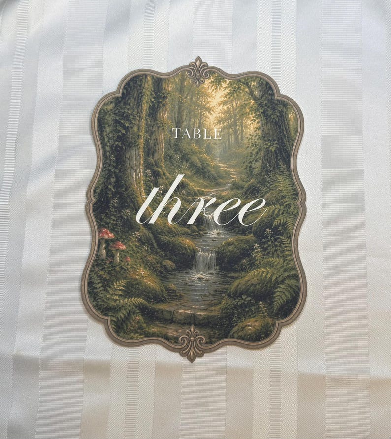 May include: A scalloped-edge table number card featuring a forest landscape with a stream, mossy rocks, and mushrooms. The text "TABLE" appears in a serif font above the word "three" written in a white cursive script centered on the card.