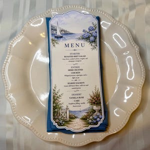 May include: A rectangular menu card with a scalloped edge, featuring a coastal illustration of a sailboat and floral garden. The text lists a starter of roasted beet salad, entrees of herb crusted chicken or seared salmon, and a vanilla rose cake dessert.