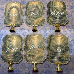 May include: Six scalloped-edge table number cards featuring forest illustrations and gold-toned metal stands. Each card displays the word TABLE followed by the written number one through six in white script, set against scenes of trees, paths, a cottage, a deer, and a stone bridge.