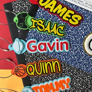 May include: Five personalized plastic bag tags with metal wire loops. The tags feature the names James, Isaac, Gavin, Quinn, and Tommy in raised, colorful lettering, each accompanied by a small icon: a pizza slice, game controller, planet, basketball, and soccer ball.