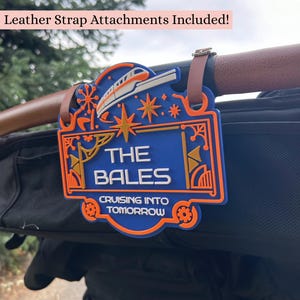 May include: A blue and orange rectangular sign with a monorail graphic, star accents, and the text THE BALES CRUISING INTO TOMORROW. The sign features two brown leather strap attachments at the top for securing it to a handle or bar.