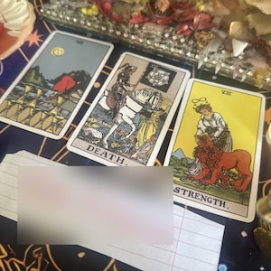 May include: Three tarot cards arranged on a dark surface. The left card shows a figure walking away from eight cups. The center card, labeled DEATH, depicts a skeleton on a white horse. The right card, labeled STRENGTH, shows a person with a lion.
