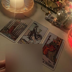 Puede incluir: Una vela encendida sobre un platillo color melocot&oacute;n junto a tres cartas del tarot. Las cartas presentan ilustraciones de figuras y las palabras "KN" y "of WANDS". Una mano sostiene una tarjeta en blanco.