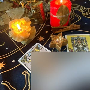 Puede incluir: Una escena m&iacute;stica con velas encendidas, cartas del tarot y objetos decorativos. Se ve una vela roja con una hoja y cuerda, una vela en forma de estrella y una vela en forma de flor. La escena est&aacute; ambientada sobre una tela azul oscuro con patrones celestiales dorados.
