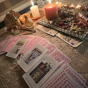 Op de afbeelding: Een uitgespreide set tarotkaarten met roze patronen op de achterkant en Turkse tekst op de voorzijde, waaronder Kılı&ccedil; D&ouml;rtl&uuml;s&uuml;, Kılı&ccedil; Kralı en Tılısım Beşlisi. De sc&egrave;ne bevat brandende kaarsen, een glazen dienblad met gedroogde planten en biljetten van 100 euro.