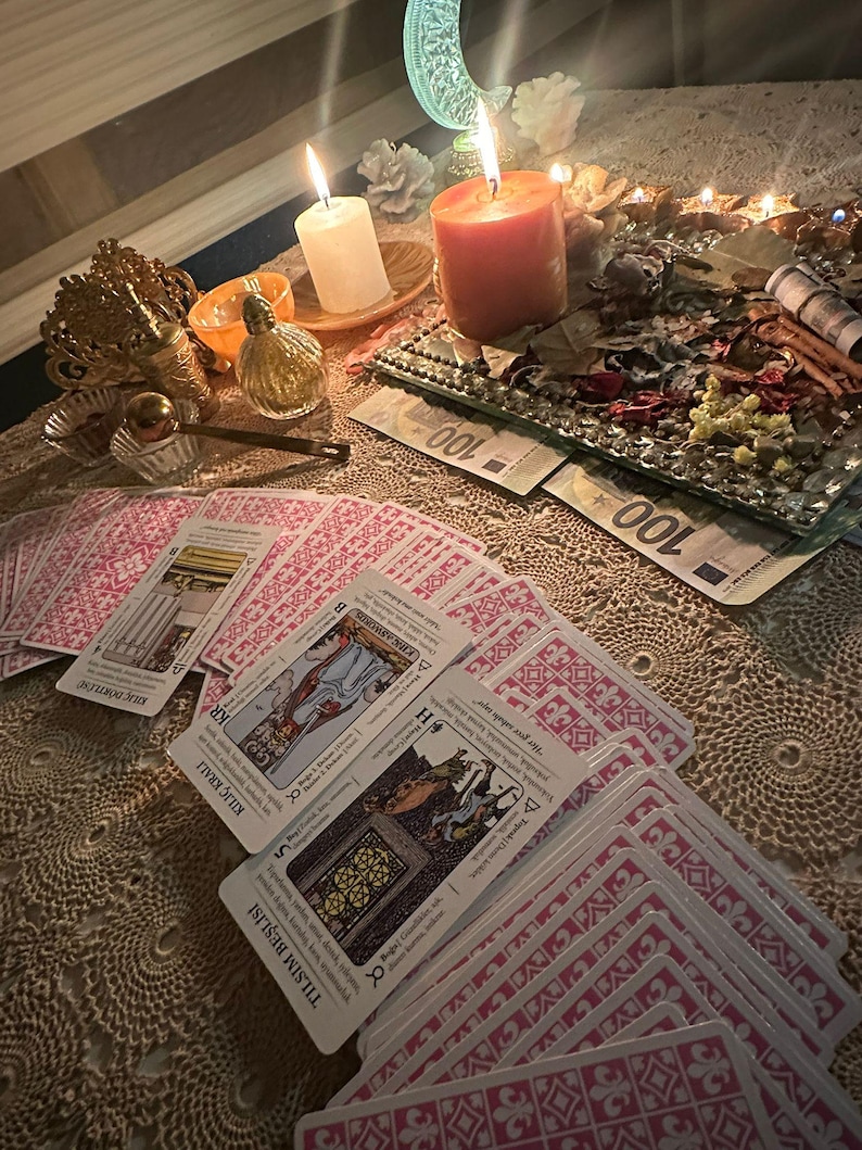 Op de afbeelding: Een uitgespreide set tarotkaarten met roze patronen op de achterkant en Turkse tekst op de voorzijde, waaronder Kılı&ccedil; D&ouml;rtl&uuml;s&uuml;, Kılı&ccedil; Kralı en Tılısım Beşlisi. De sc&egrave;ne bevat brandende kaarsen, een glazen dienblad met gedroogde planten en biljetten van 100 euro.
