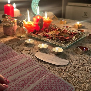 Op de afbeelding: Een set tarotkaarten met roze en witte patronen op de achterkant, een glazen dienblad gevuld met gedroogde rode rozenblaadjes, meerdere brandende rode en witte kaarsen, een klein glazen parfumflesje en een halve maan ornament op een gehaakt kanten kleed.