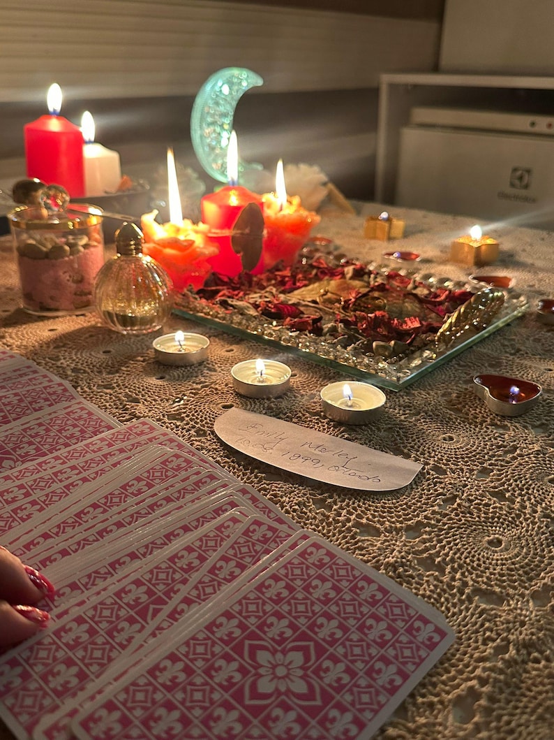 Op de afbeelding: Een set tarotkaarten met roze en witte patronen op de achterkant, een glazen dienblad gevuld met gedroogde rode rozenblaadjes, meerdere brandende rode en witte kaarsen, een klein glazen parfumflesje en een halve maan ornament op een gehaakt kanten kleed.