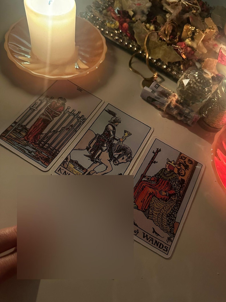 Op de afbeelding: Een brandende kaars op een perzikvormige schotel naast drie tarotkaarten. De kaarten bevatten illustraties van figuren en de woorden "KN" en "of WANDS". Een hand houdt een blanco kaart vast.