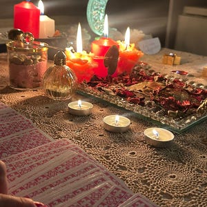Op de afbeelding: Een set tarotkaarten met een rood-wit patroon op de achterkant, uitgespreid op een beige kanten tafelkleed. De sc&egrave;ne bevat verschillende brandende rode en witte kaarsen, een glazen pot met roze inhoud, een klein parfumflesje en een halve maan ornament.