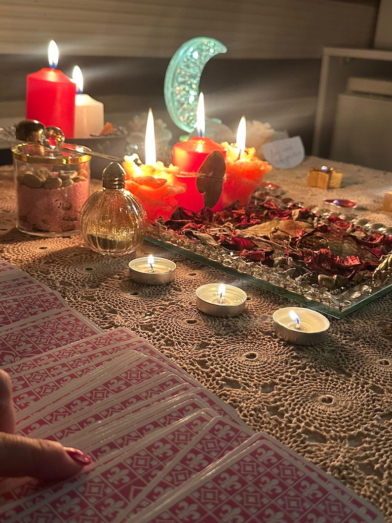 Op de afbeelding: Een set tarotkaarten met een rood-wit patroon op de achterkant, uitgespreid op een beige kanten tafelkleed. De sc&egrave;ne bevat verschillende brandende rode en witte kaarsen, een glazen pot met roze inhoud, een klein parfumflesje en een halve maan ornament.