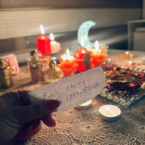 Op de afbeelding: Een hand houdt een wit rechthoekig papier vast met de handgeschreven tekst Emily Morley 1999, Orlando. Op de achtergrond branden meerdere rode en witte kaarsen, waaronder een halvemaanvormig licht, naast kleine glazen flesjes en een dienblad.