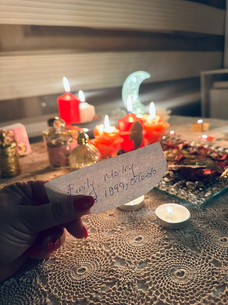 Op de afbeelding: Een hand houdt een wit rechthoekig papier vast met de handgeschreven tekst Emily Morley 1999, Orlando. Op de achtergrond branden meerdere rode en witte kaarsen, waaronder een halvemaanvormig licht, naast kleine glazen flesjes en een dienblad.