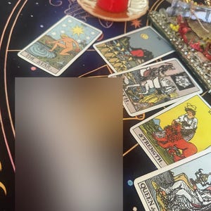 Puede incluir: Varias cartas del tarot, incluyendo "La Estrella", "Fuerza" y "Reina de Copas", se encuentran sobre una superficie oscura y estampada. Una vela roja encendida y una bandeja decorativa completan la escena, evocando un tema espiritual o de adivinaci&oacute;n.