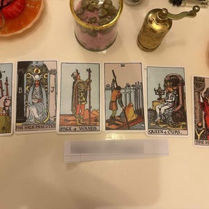 May include: A spread of six tarot cards arranged horizontally, featuring the Six of Wands, The High Priestess, Page of Wands, Six of Swords, Queen of Cups, and The Hierophant. Each card displays detailed illustrations with vibrant colors and text.