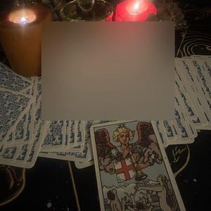 Op de afbeelding: Een tarotkaart met het woord "JUDGEMENT" onderaan, met een engel die op een trompet blaast. De kaart wordt omringd door andere tarotkaarten, brandende kaarsen en decoratieve items, wat een mystieke sfeer cre&euml;ert.
