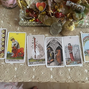 May include: A set of six tarot cards arranged in a row, including the Eight of Pentacles, Strength, Ace of Wands, Three of Pentacles, Eight of Swords, and Ten of Cups, featuring various illustrations of figures, symbols, and landscapes.