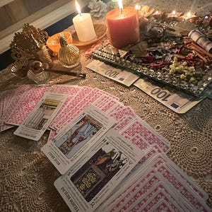 Op de afbeelding: Een mystieke sc&egrave;ne met tarotkaarten met roze achterkanten en illustraties, verspreid op een kanten tafelkleed. Brandende kaarsen, een decoratief dienblad met gedroogde bloemen en valuta cre&euml;ren een sfeer van waarzeggerij en spiritualiteit.