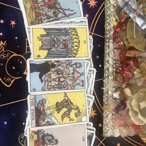 May include: A collection of tarot cards arranged in a vertical row, including the Five of Wands, Nine of Cups, Seven of Cups, The Fool, and Eight of Pentacles. The cards feature colorful illustrations and Roman numerals, displayed on a dark blue cloth.