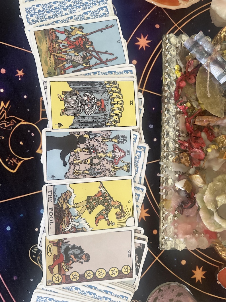 May include: A collection of tarot cards arranged in a vertical row, including the Five of Wands, Nine of Cups, Seven of Cups, The Fool, and Eight of Pentacles. The cards feature colorful illustrations and Roman numerals, displayed on a dark blue cloth.