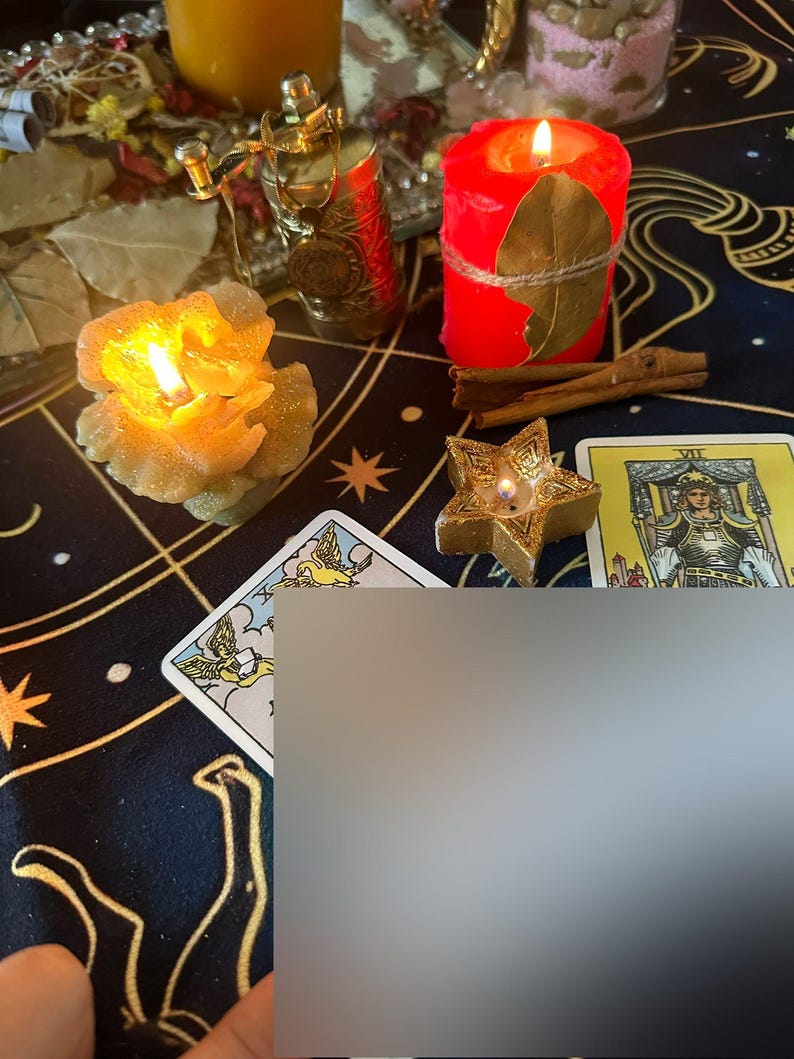 Op de afbeelding: Een mystieke sc&egrave;ne met brandende kaarsen, tarotkaarten en decoratieve items op een patroonstof. Een rode kaars met een blad en touw, een stervormige kaars en een bloemvormige kaars zijn zichtbaar. Kaneelstokjes en een gouden parfumflesje zijn ook aanwezig.