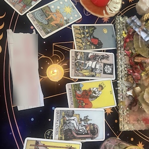May include: A collection of tarot cards arranged on a dark surface with celestial patterns. Visible cards include The Star, Death, Strength, Queen of Cups, and Four of Wands. A red candle in a glass holder, a crystal tray, and a small jar are also present.