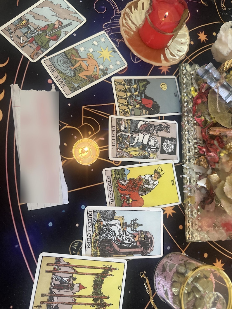 May include: A collection of tarot cards arranged on a dark surface with celestial patterns. Visible cards include The Star, Death, Strength, Queen of Cups, and Four of Wands. A red candle in a glass holder, a crystal tray, and a small jar are also present.
