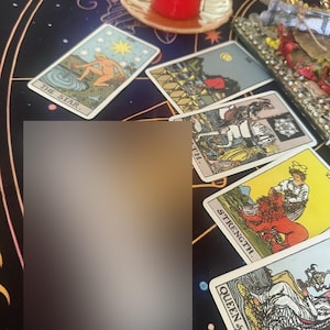 Op de afbeelding: Een verzameling tarotkaarten, waaronder "De Ster", "Kracht" en "Koningin van Bekers", ligt op een donkere, bedrukte doek. Een brandende rode kaars en een decoratief dienblad zijn ook zichtbaar.