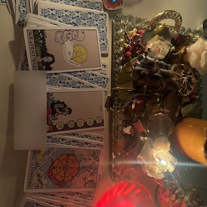 Op de afbeelding: Een verzameling tarotkaarten met blauwe patronen op de achterkant en verschillende kaartillustraties. De kaart 'Aas van Pentakels' is zichtbaar, samen met andere kaarten. De sc&egrave;ne bevat kaarsen, kristallen en decoratieve elementen, wat een mystieke sfeer cre&euml;ert.