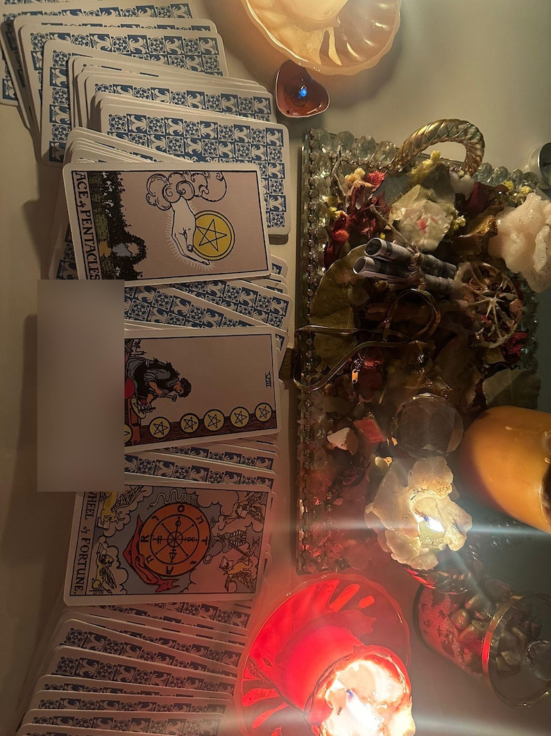 Op de afbeelding: Een verzameling tarotkaarten met blauwe patronen op de achterkant en verschillende kaartillustraties. De kaart 'Aas van Pentakels' is zichtbaar, samen met andere kaarten. De sc&egrave;ne bevat kaarsen, kristallen en decoratieve elementen, wat een mystieke sfeer cre&euml;ert.
