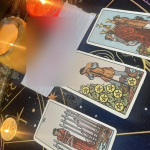 May include: Three tarot cards arranged on a dark surface with gold celestial patterns. The cards include the Three of Cups, the Seven of Pentacles, and the Eight of Swords. Two lit candles with orange and yellow flames are positioned nearby.
