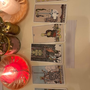 May include: A vertical arrangement of six tarot cards including the Six of Pentacles, Ace of Swords, Four of Pentacles, Five of Pentacles, Queen of Swords, and The Star. To the left are two lit candles in gold-toned holders and a glass jar with pink sand.