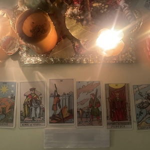 May include: A row of six tarot cards arranged horizontally, featuring The Star, King of Cups, Six of Swords, Five of Swords, Justice, and Knight of Swords. The cards are illuminated by warm, bright candlelight from above, creating a glowing effect.