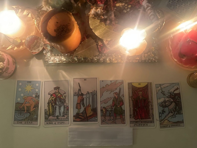 May include: A row of six tarot cards arranged horizontally, featuring The Star, King of Cups, Six of Swords, Five of Swords, Justice, and Knight of Swords. The cards are illuminated by warm, bright candlelight from above, creating a glowing effect.