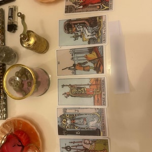 May include: A vertical spread of six tarot cards featuring The Hierophant, Queen of Cups, Six of Swords, Page of Wands, The High Priestess, and Six of Wands. Beside the cards are a brass hand mill, a glass jar with pink contents, and two small dishes.