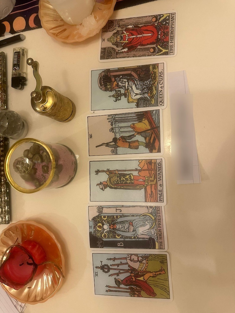 May include: A vertical spread of six tarot cards featuring The Hierophant, Queen of Cups, Six of Swords, Page of Wands, The High Priestess, and Six of Wands. Beside the cards are a brass hand mill, a glass jar with pink contents, and two small dishes.