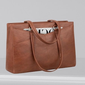 May include: A brown leather tote bag with two handles and a front pocket. The bag has a rectangular shape and a structured design. A magazine is visible inside the bag. The bag is sitting on a white surface.